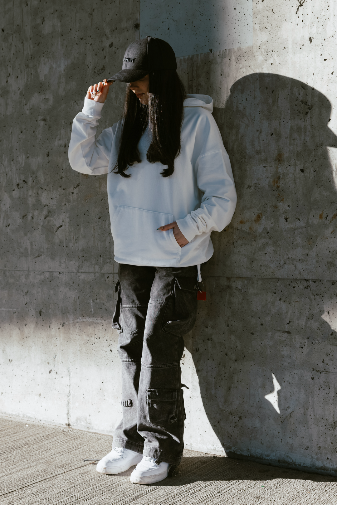 Women in white hoodie and black hat in streetwear outfit infront on concrete wall
