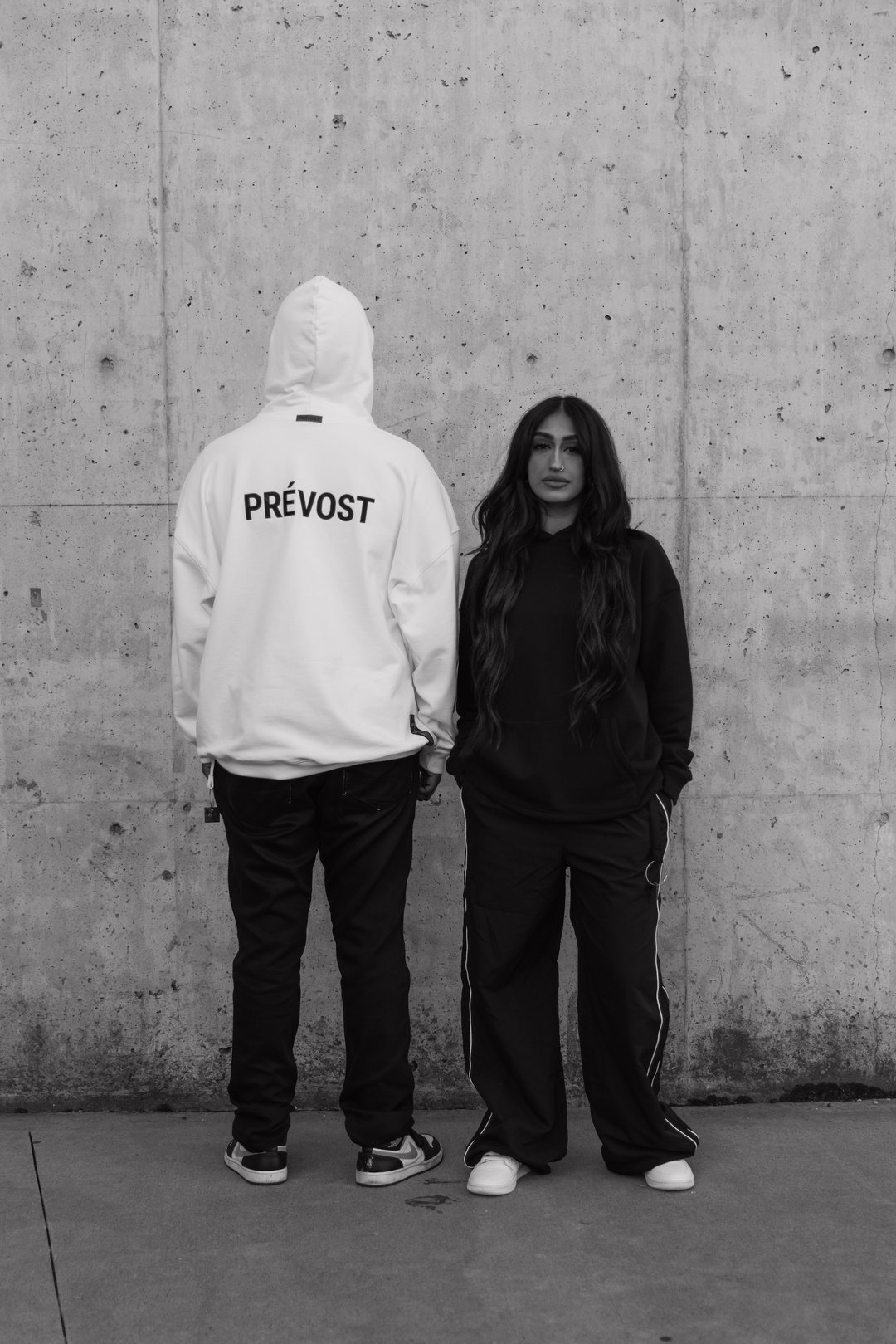 Man and women standing side by side with white and black hoodie in front of wall